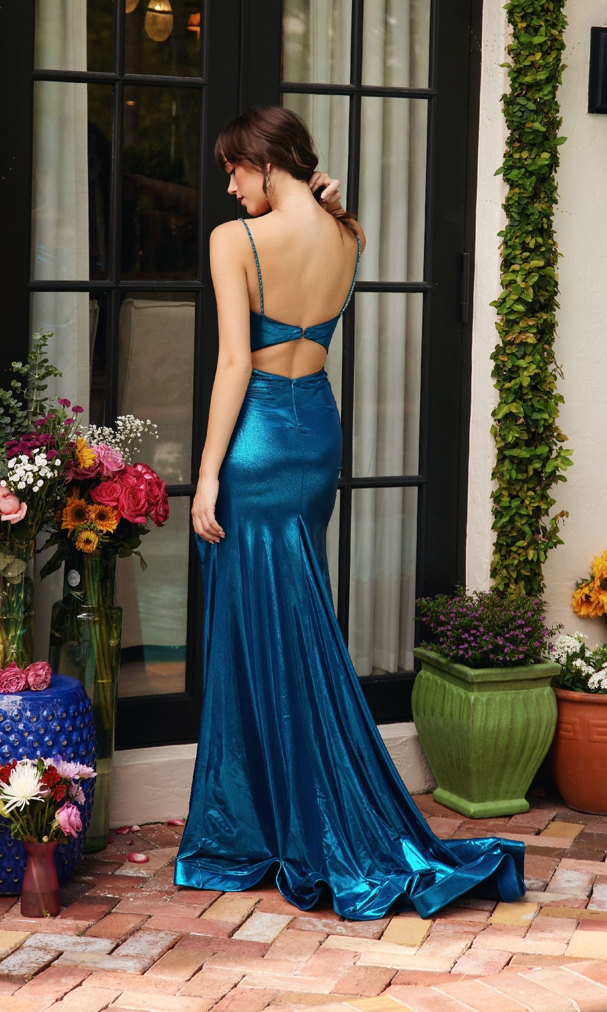 A woman in the Ava Presley 40101 long cowl-neck prom dress, featuring a shiny teal metallic finish and side slit, stands on a brick patio near potted plants and flowers by a glass door.